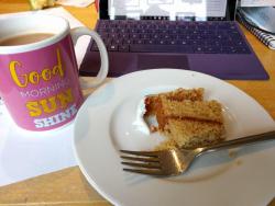 Image of cup of tea, and a piece of cake on a plate, with a computer open in the background